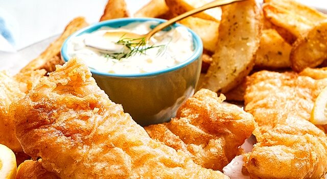 cream coloured oval platter with beer battered fish, French fries, lemon wedges and tartar sauce in a side bowl.