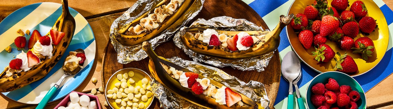 Berry shortcake banana boats on picnic table with plate of strawberries and raspberries to the side.
