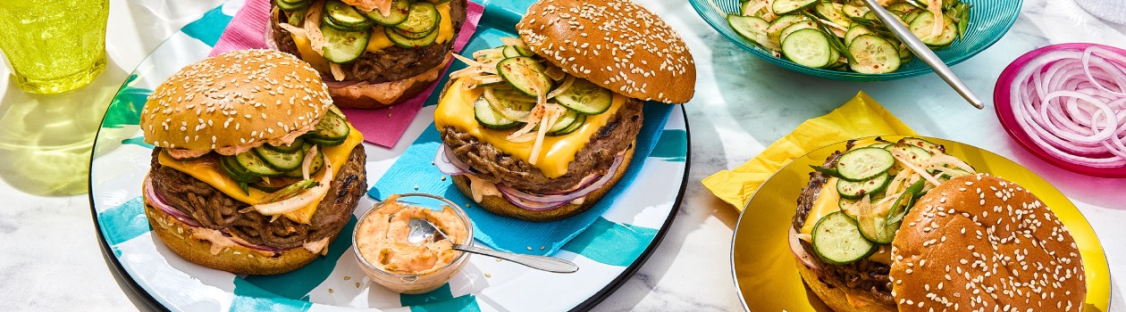 Three big Korean-style burgers and kimchi mayo on a blue and white striped plate, a burger on a yellow plate and Korean-style pickled cucumber salad in a blue serving bowl on a marbled surface.
