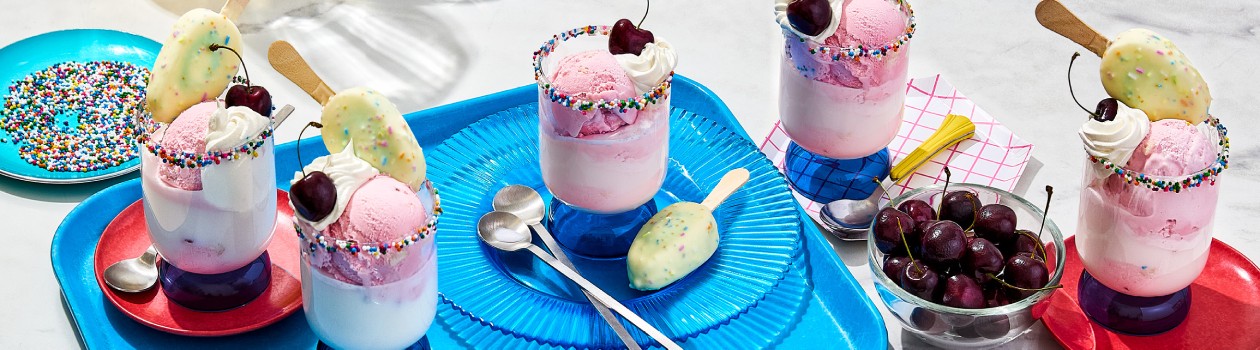 Five cups of the Birthday Cake Mini Bar Float with cherries in a bowl off to the side, spoons, a birthday cake mini bar, sprinkles, and various plates.