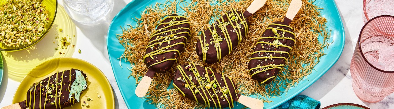 Four Dubai Inspired Chocolate Pistachio Bars on a bed of kataifi with one bar off to the side with a bite taken out and a bowl of pistachios.