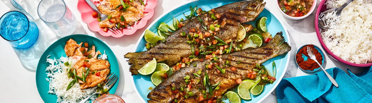 A plate with two whole trouts and the relish with sliced lime, two smaller plates with the cooked fish on rice, a small bowl of relish and one of chili powder and one larger bowl of white rice.