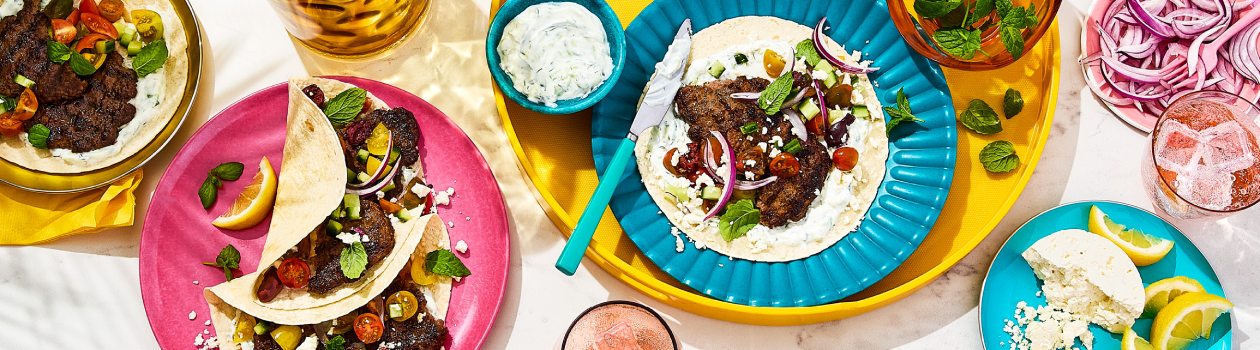 Overhead shot of four Greek-inspired smashed burger tacos with tzatziki on the side.
