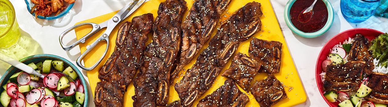 Overhead shot of kalbi Korean-style ribs on a yellow platter with scissors and a bowl of fresh salad on the side.