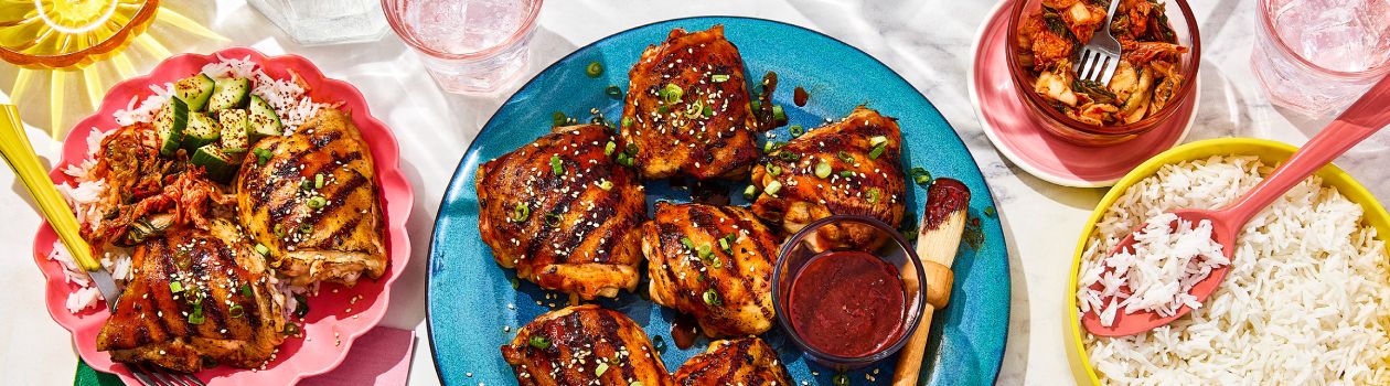 Two plates of Korean-style BBQ chicken, a bowl of kimchi and a bowl of rice on a marbled surface.
