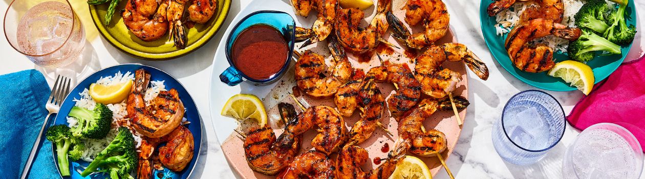 Overhead shot of four plates of Korean-style fiery shrimp skewers, with accompanying sides including rice, broccoli and lemon wedges.