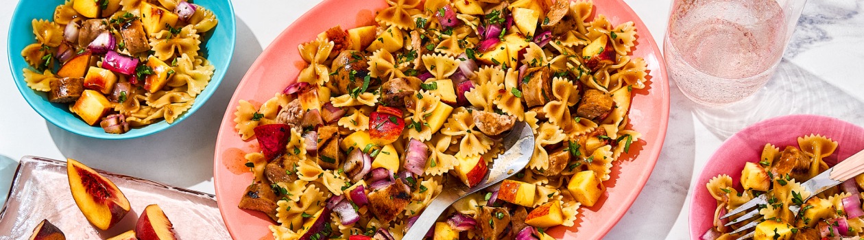 Platter filled with bowtie pasta salad and fresh peaches, with two bowls with the same pasta to the side.