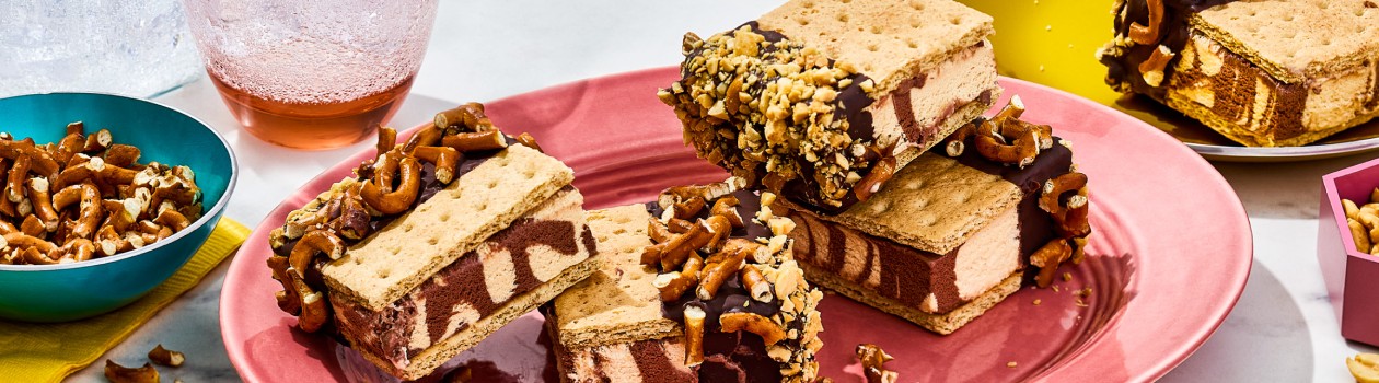 Four Peanut Butter S’mores Ice Cream Sandwiches on a plate with a small bowl of chopped pretzels off to the side and another plate with a single sandwich.