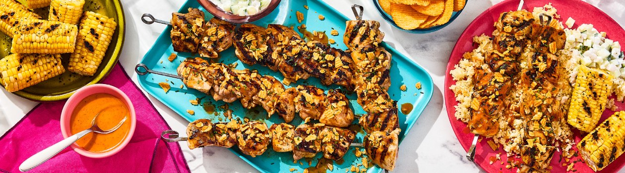A platter of four Portuguese-Inspired Piri Piri Hot Honey BBQ Chicken Kabobs with a sauce, a small plate with more skewers, rice, and corn, and chips, corn, and more sauce in bowls off to the side.