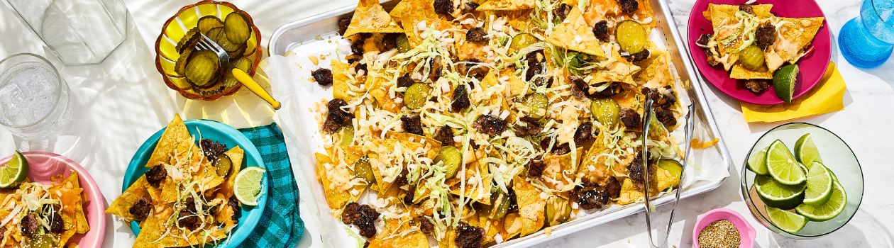 Smashed burger nachos on a sheet pan and small plates, with small bowls of pickle slices and limes on the side.