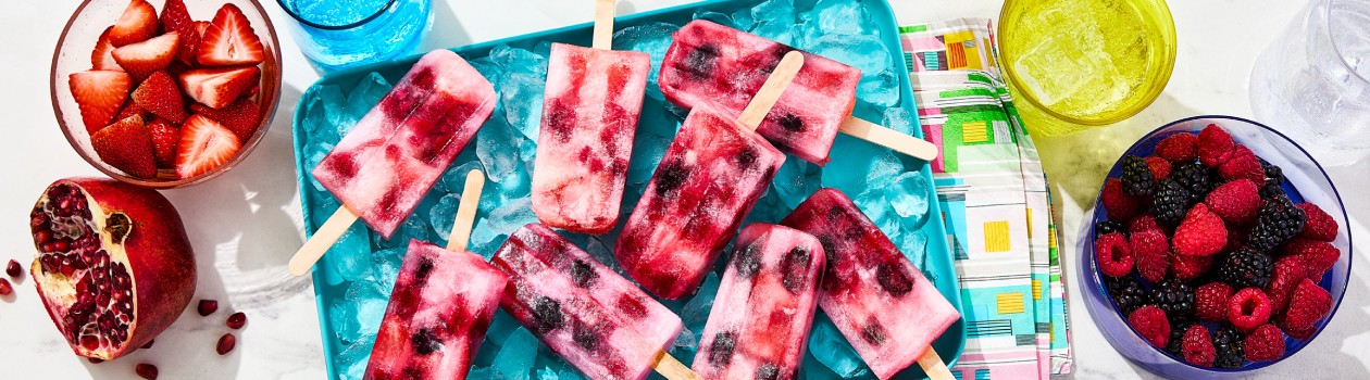 A platter filled with ice and several Sparkling Water Popsicles lying on it with bowls of berries and half a pomegranate off to the side.
