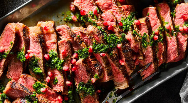 Grilled Syrian Seven Spice Rib Steak sliced on a grey platter with a nearby bowl full of green, herbaceous Chermoula and a bowl of grilled corn.